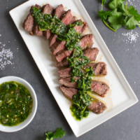 Slices of steak arranged on a white plate drizzled with cilantro chimichurri, with cilantro and salt flakes surrounding on a slate gray background.