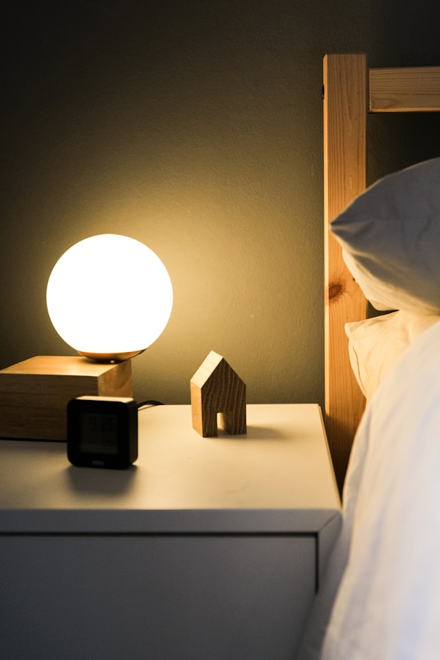 An orange-tinted nightlight in a dark room on a white nightstand next to a bed with white sheets.