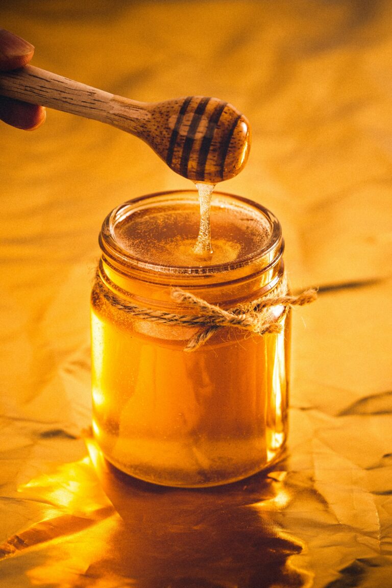 A drizzle of honey pouring into a clear jar of honey, backlit by a golden glow.