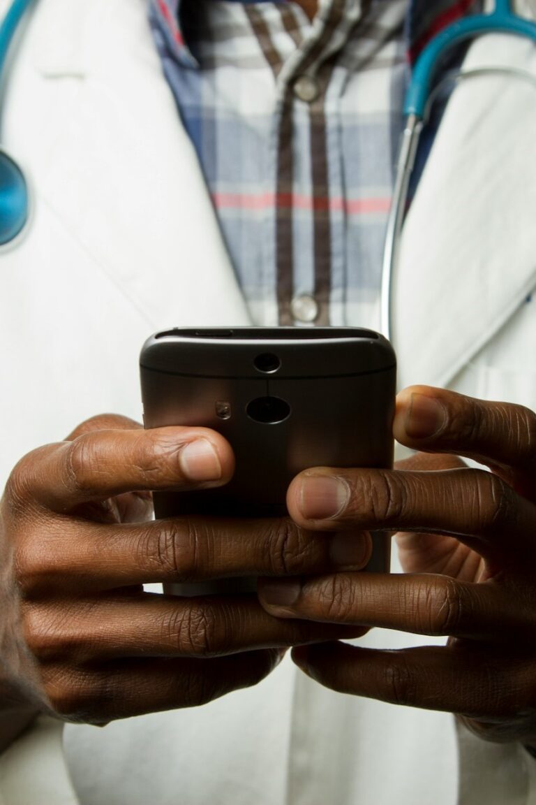 A Black doctor in a white lab coat with a stethoscope over his shoulders holding a cell phone as if he is looking something up or taking notes.