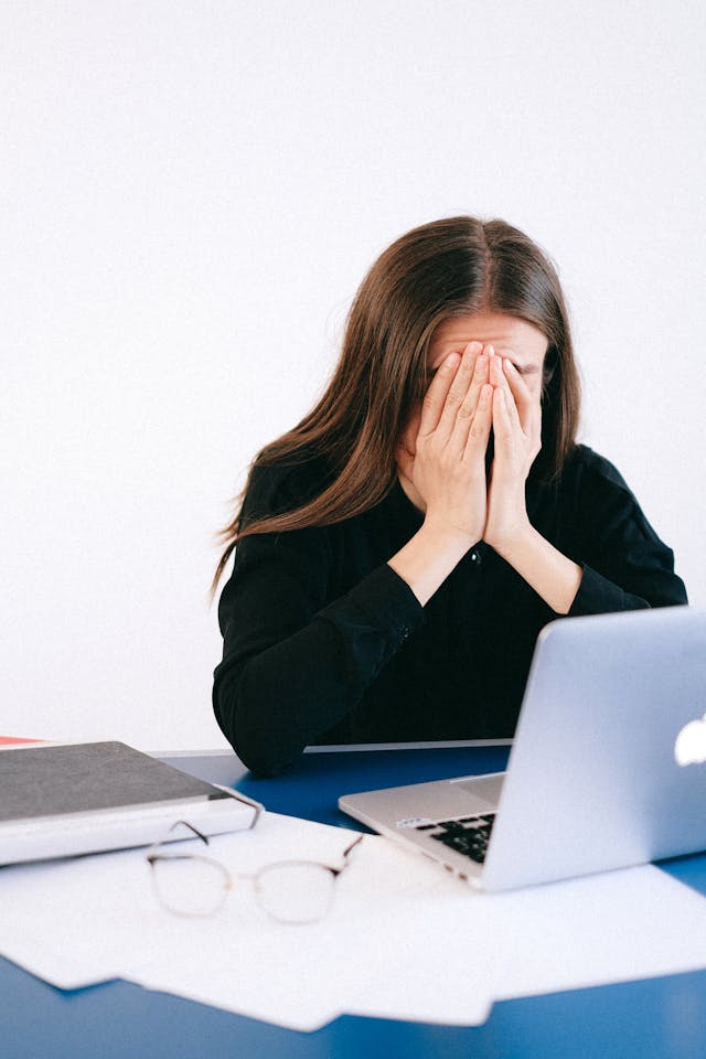 A brunette woman in a black sweater sitting in front of a macbook with her face buried in her hands.